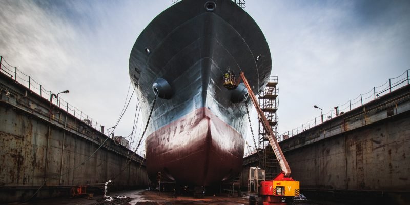 A dramatic low-angle shot of the massive bow of a bulk carrier in drydock, showcasing the imposing scale of the vessel as it towers above. The ship’s hull is exposed, with scaffolding, a cherry picker and maintenance equipment visible around the drydock area. The anchor and anchor chain of the vessel lie in front of it. A team of workers is painting the hull under a dramatic cloudy sky. The image captures the engineering marvel of large-scale shipbuilding and repair, ideal for themes of maritime industry, ship maintenance, heavy machinery, and industrial work